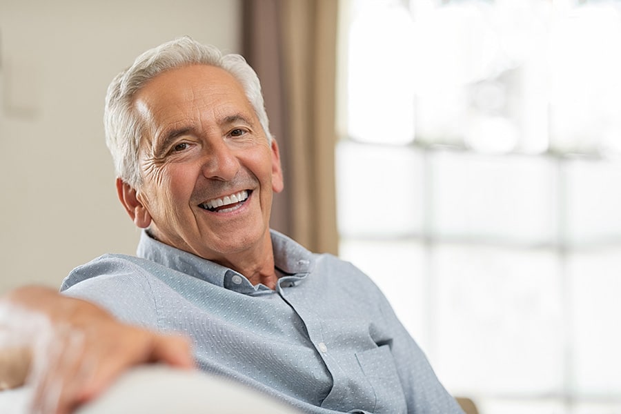 Portrait of happy senior man smiling at home. Old man relaxing on sofa and looking at camera. Portrait of elderly man enjoying retirement.